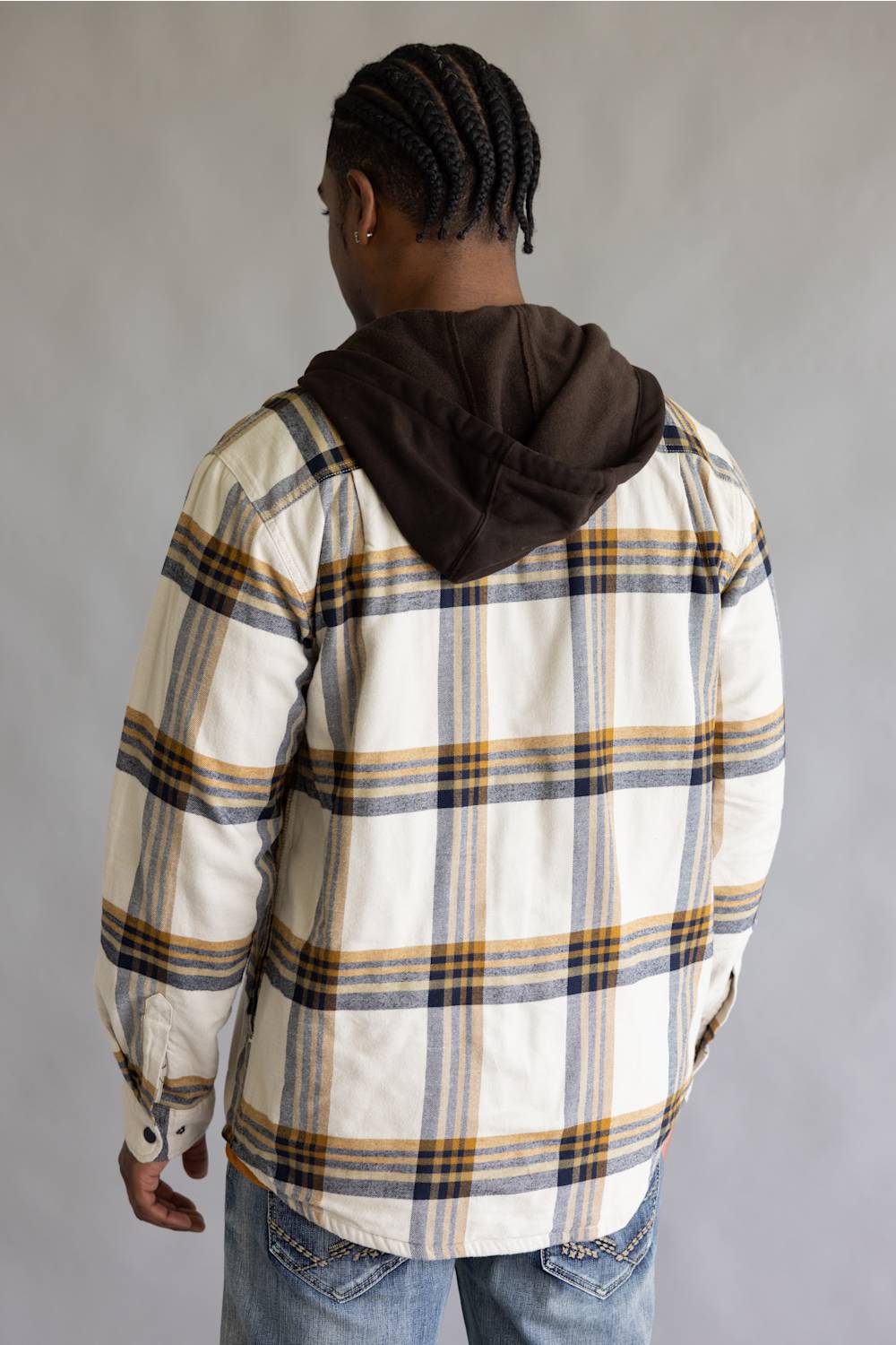 A man viewed from behind wearing a cream-colored plaid hooded overshirt with yellow and navy stripes, paired with blue jeans, against a plain gray background.