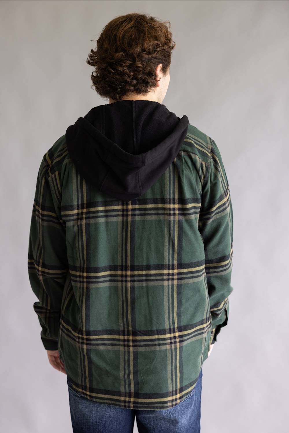 A person viewed from the back wearing a green plaid hooded jacket with a black hood, paired with blue jeans, against a plain gray studio background.