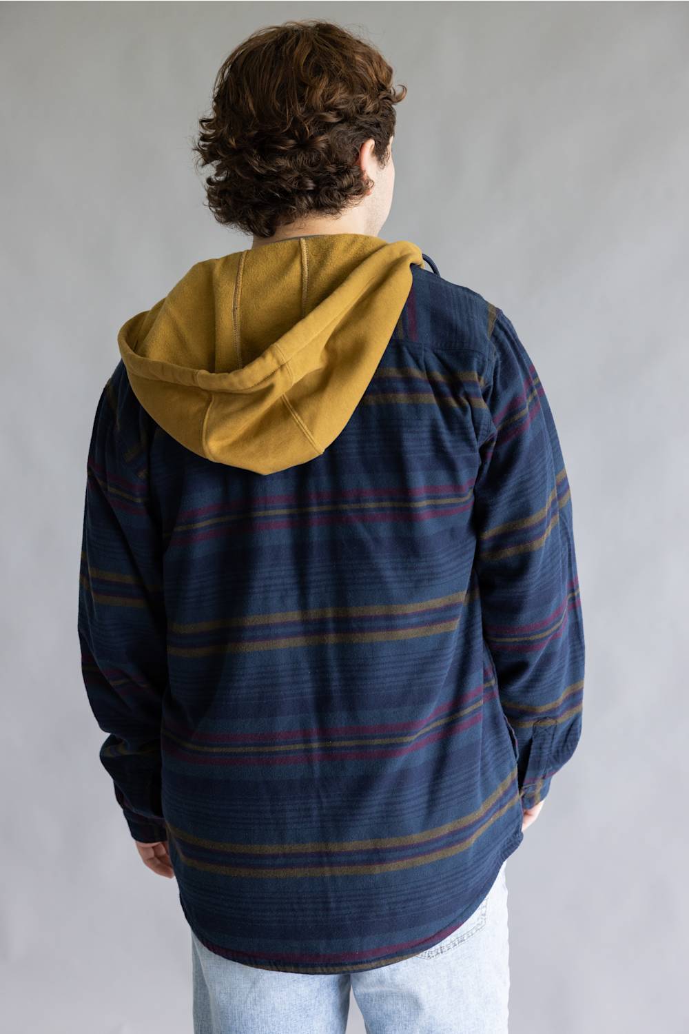 A person shown from the back wearing a navy blue striped hooded shirt-jacket with a mustard yellow hood, paired with light blue jeans, against a plain gray studio background.