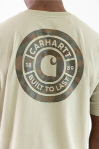 Close-up of the back of a person wearing a light beige Carhartt T-shirt with a large circular camouflage graphic and the words CARHARTT BUILT TO LAST printed in white.