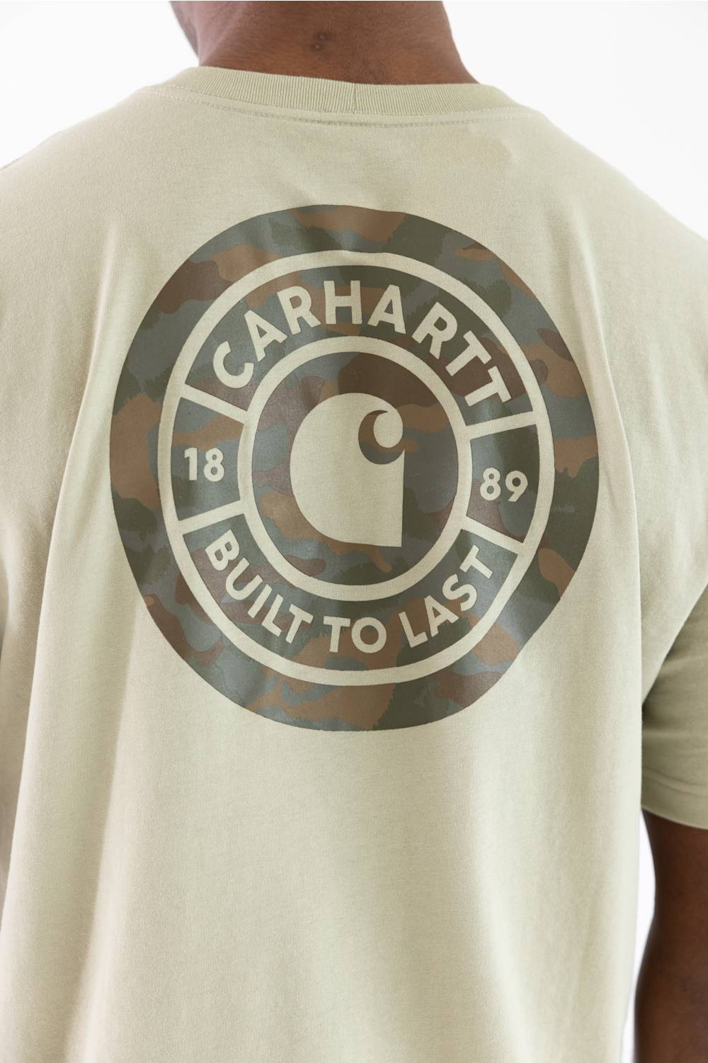 Close-up of the back of a person wearing a light beige Carhartt T-shirt with a large circular camouflage graphic and the words CARHARTT BUILT TO LAST printed in white.