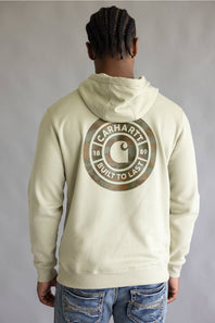 A man viewed from behind wearing a cream-colored Carhartt hoodie with a large brown camouflage circular logo on the back, paired with blue jeans against a neutral gray background.