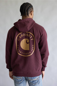A person shown from the back wearing a maroon hooded sweatshirt with a large gold Carhartt logo graphic on the back, paired with blue jeans against a plain gray studio background.