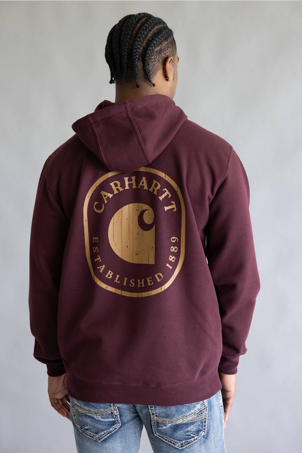 A person shown from the back wearing a maroon hooded sweatshirt with a large gold Carhartt logo graphic on the back, paired with blue jeans against a plain gray studio background.