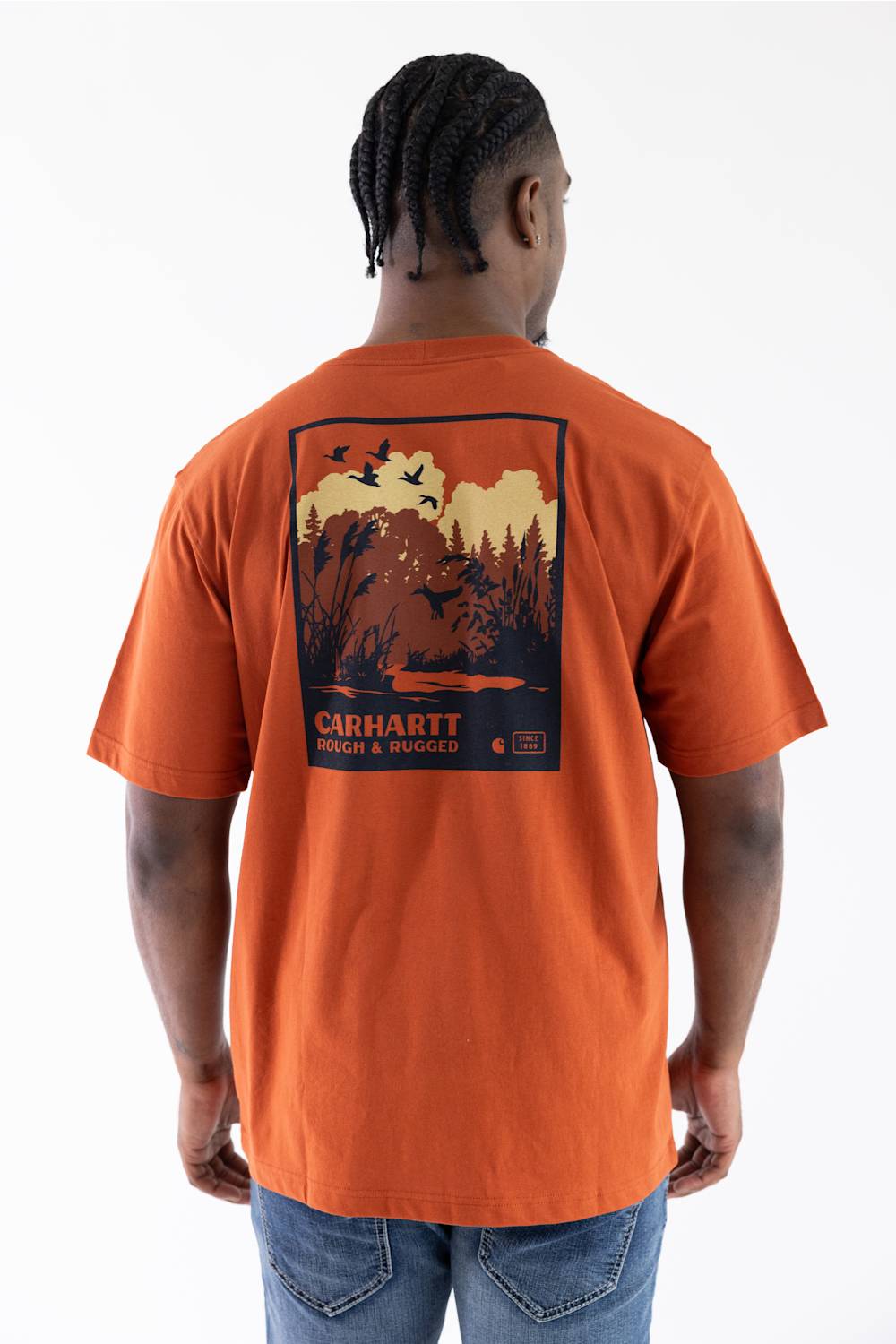 A man viewed from behind wearing a Carhartt orange short-sleeve t-shirt with a large back graphic showing a bear, trees, and the text 'CARHARTT WOOD & WOOD', paired with blue jeans against a white studio background.
