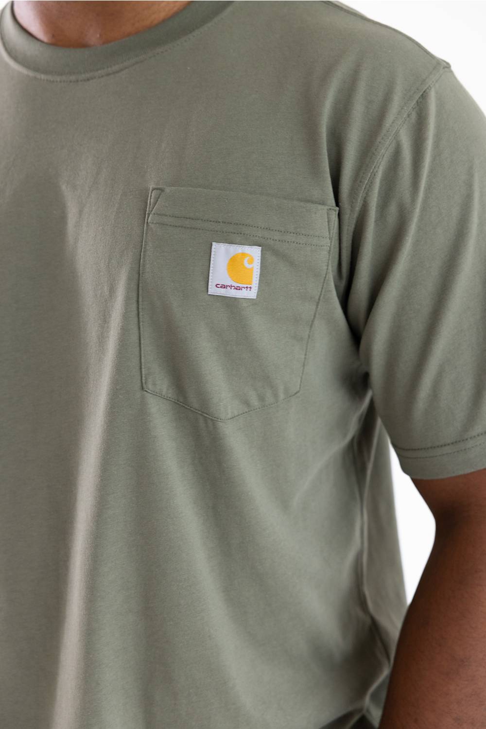 Close-up of a person wearing a muted sage green Carhartt pocket t-shirt, showing the chest pocket with the signature yellow Carhartt logo patch.