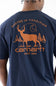 A man wearing a navy blue Carhartt t-shirt, viewed from the back. The shirt features a large orange graphic print of a deer, trees, and the Carhartt logo with the text ROOTED IN TRADITION and SINCE 1889.
