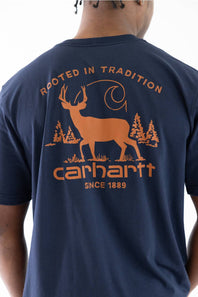 A man wearing a navy blue Carhartt t-shirt, viewed from the back. The shirt features a large orange graphic print of a deer, trees, and the Carhartt logo with the text ROOTED IN TRADITION and SINCE 1889.