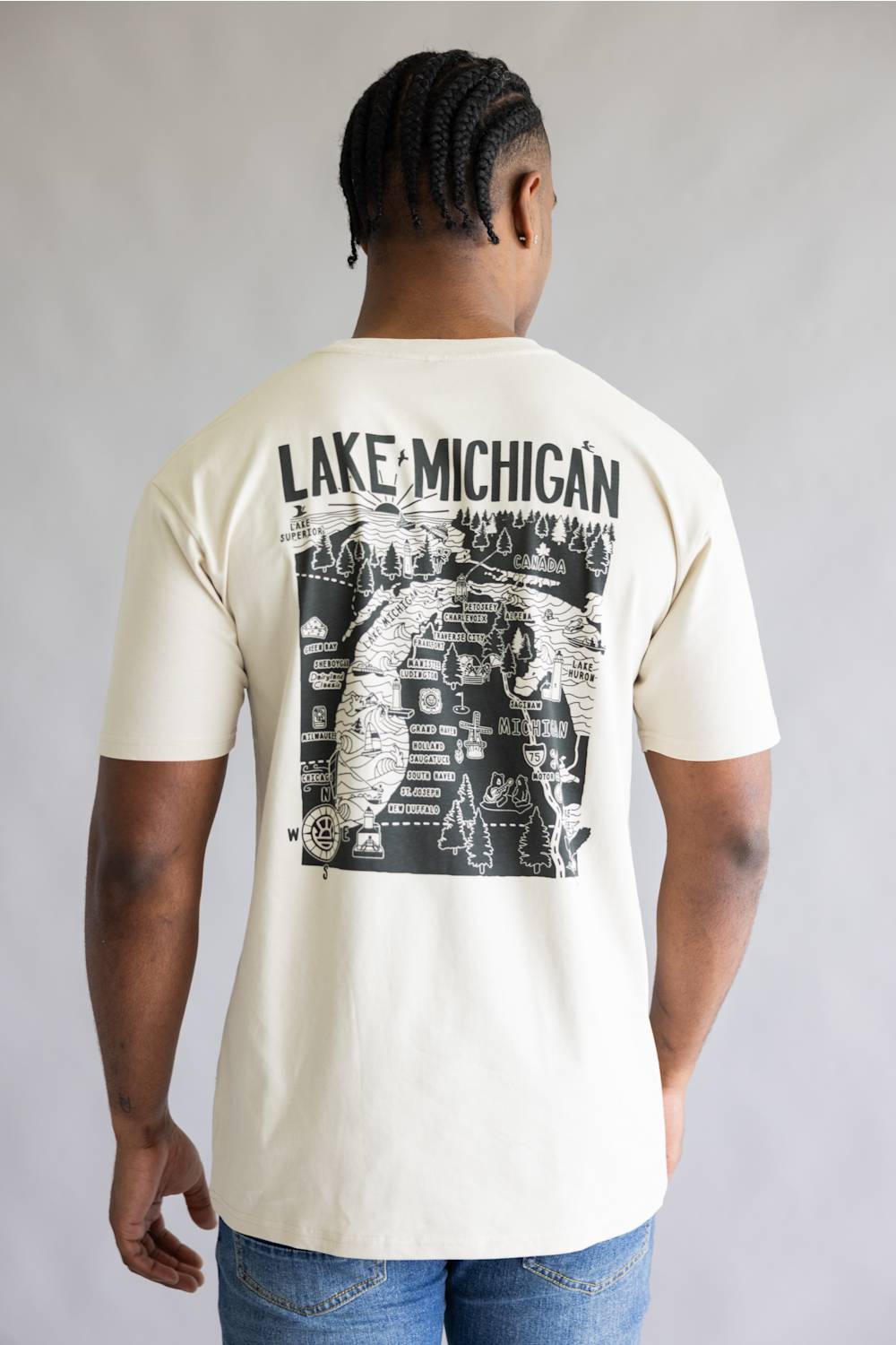A man viewed from the back wearing a cream-colored t-shirt with a large black graphic print on the back that reads 'LAKE MICHIGAN' above an illustrated map design, paired with blue jeans against a neutral gray background.