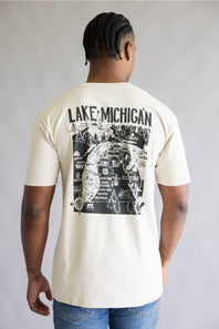 A man viewed from the back wearing a cream-colored t-shirt with a large black graphic print on the back that reads 'LAKE MICHIGAN' above an illustrated map design, paired with blue jeans against a neutral gray background.
