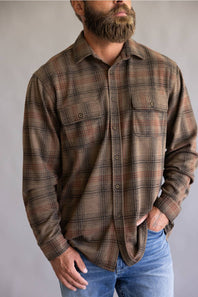 A man wearing a brown and beige plaid button-down flannel shirt with blue jeans, shown from the waist up against a neutral gray background.