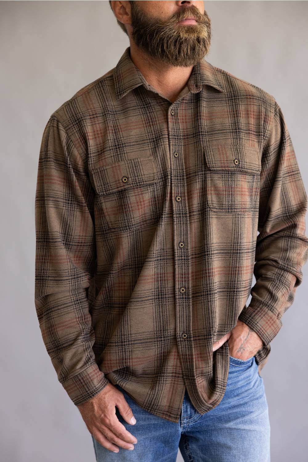 A man wearing a brown and beige plaid button-down flannel shirt with blue jeans, shown from the waist up against a neutral gray background.
