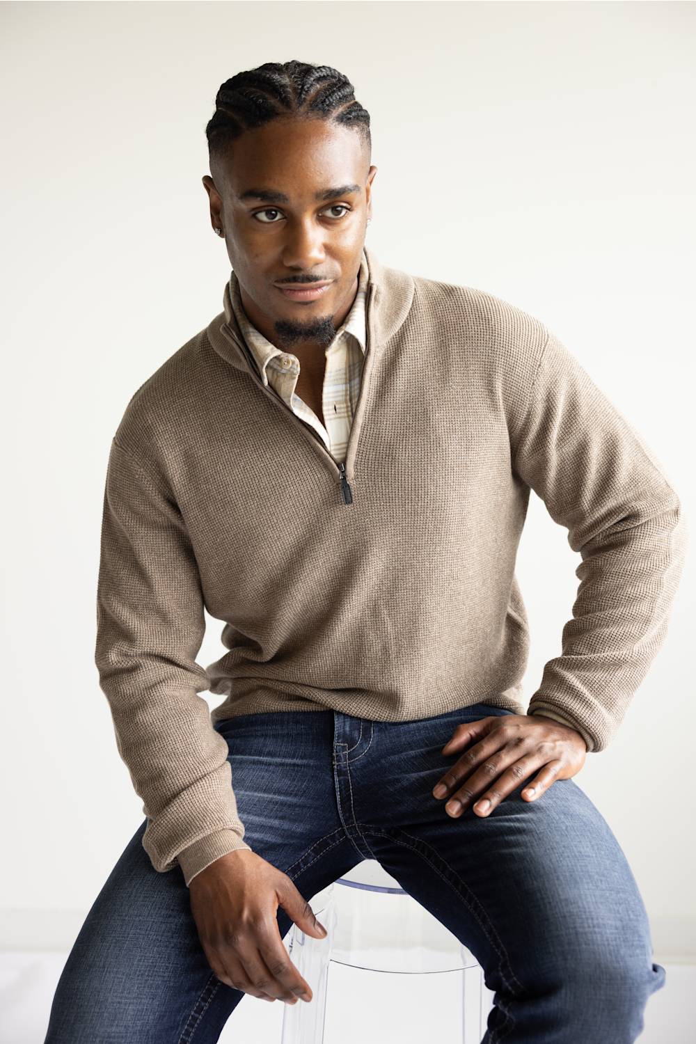 A man wearing a beige quarter-zip knit sweater over a light-colored collared shirt and blue jeans, seated against a plain light background.