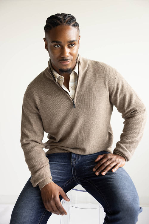 A man wearing a beige quarter-zip knit sweater over a light-colored collared shirt and blue jeans, seated against a plain light background.