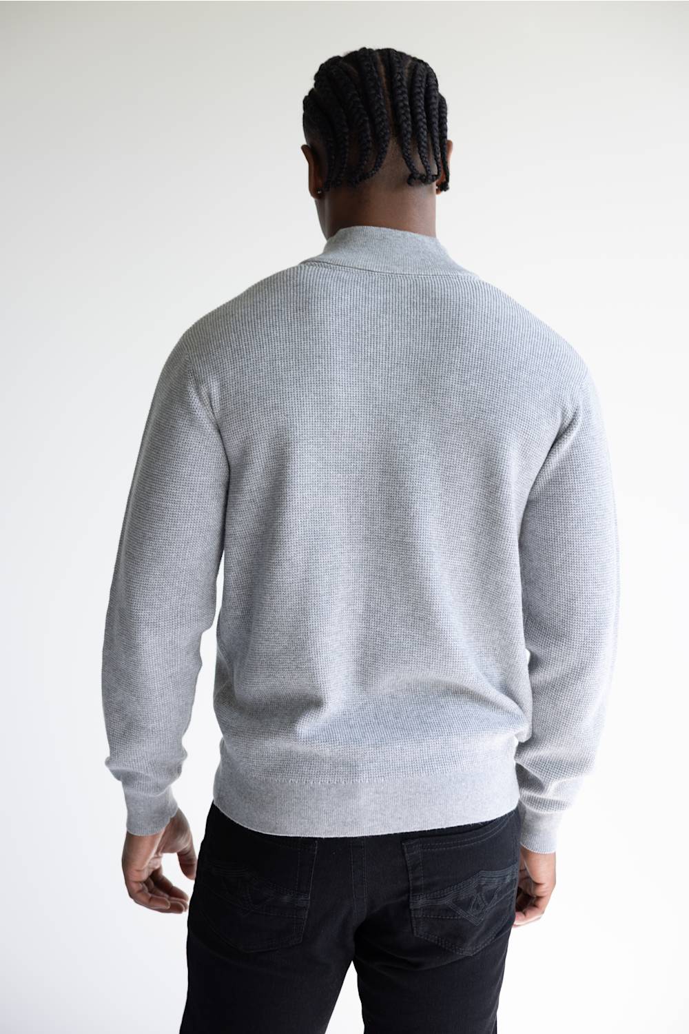A man viewed from behind wearing a light gray ribbed knit mock neck sweater and black pants, standing against a plain light gray studio background.