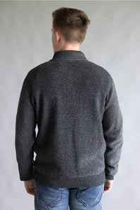 A man viewed from the back wearing a dark gray knit sweater and blue jeans, standing against a plain light gray studio background.