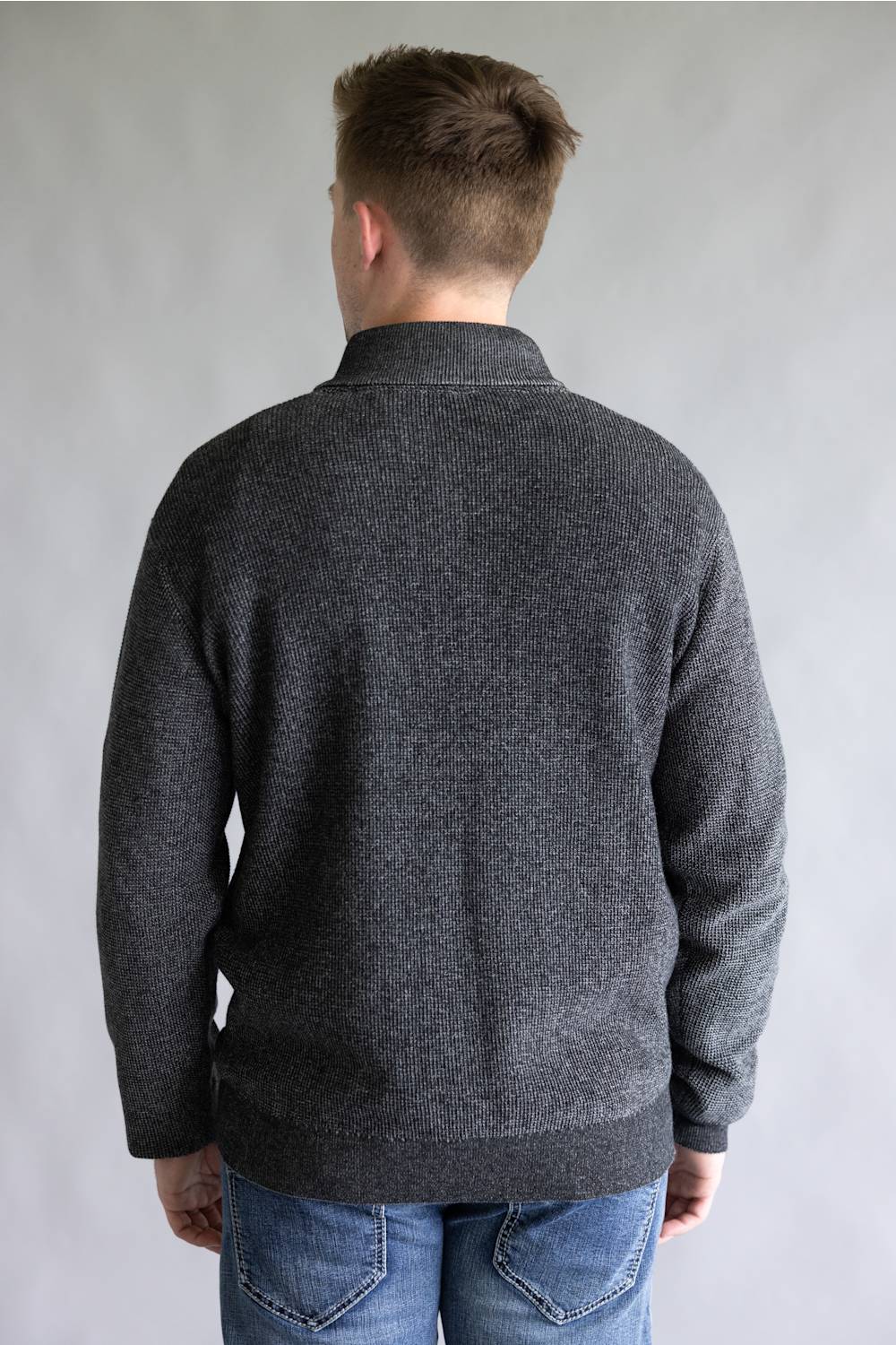 A man viewed from the back wearing a dark gray knit sweater and blue jeans, standing against a plain light gray studio background.