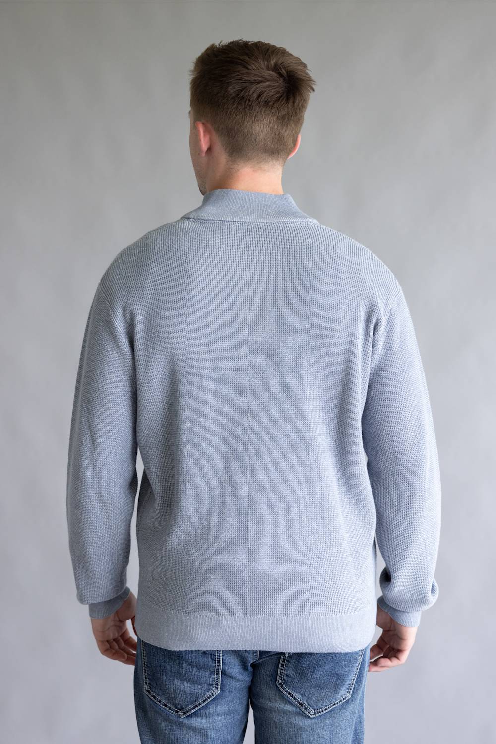 A man viewed from behind wearing a light blue ribbed knit sweater and blue jeans, standing against a neutral gray studio background.