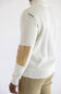 A man wearing a cream ribbed knit sweater with tan elbow patches, shown from a side-back angle against a plain white background.