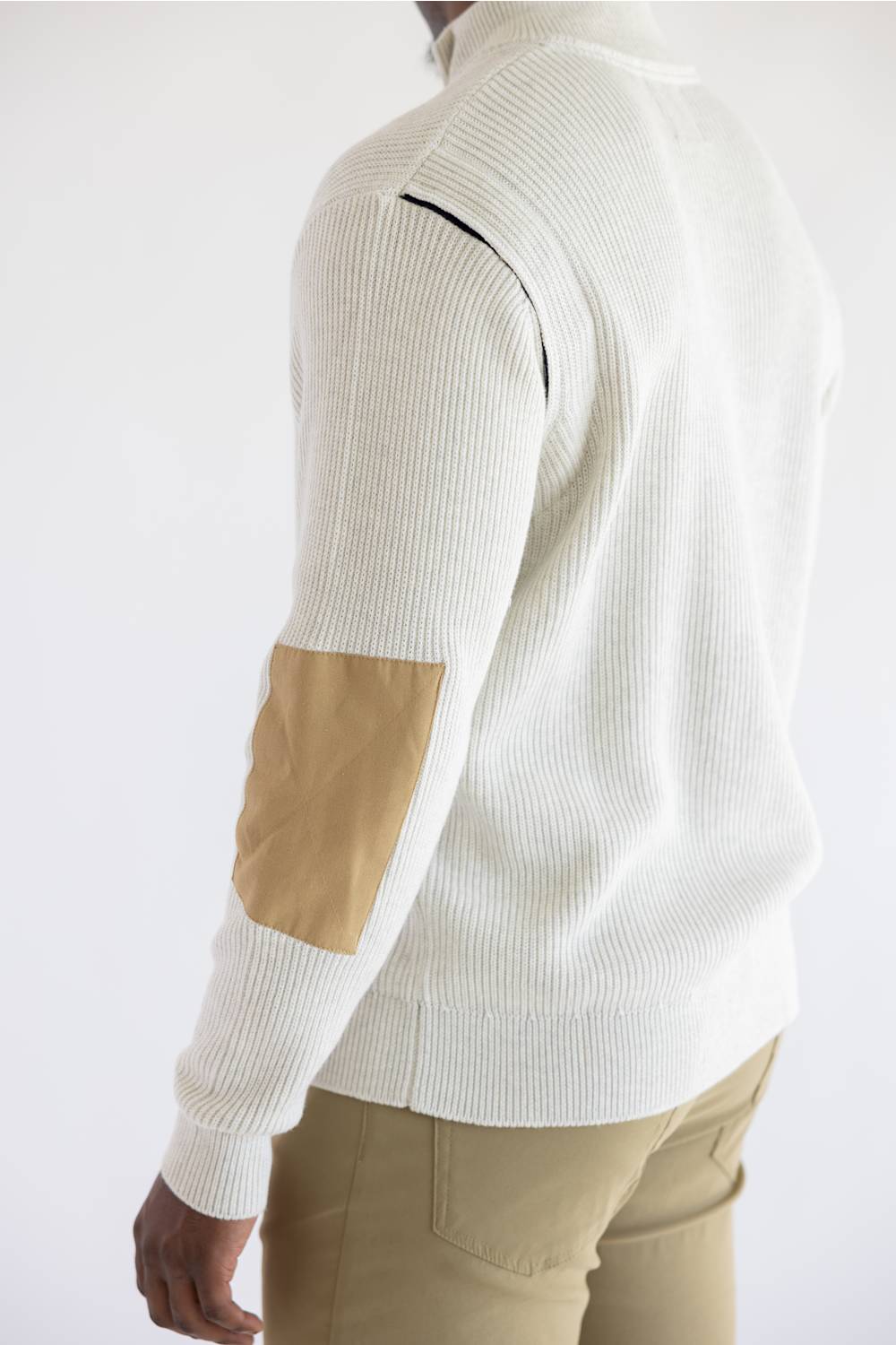 A man wearing a cream ribbed knit sweater with tan elbow patches, shown from a side-back angle against a plain white background.