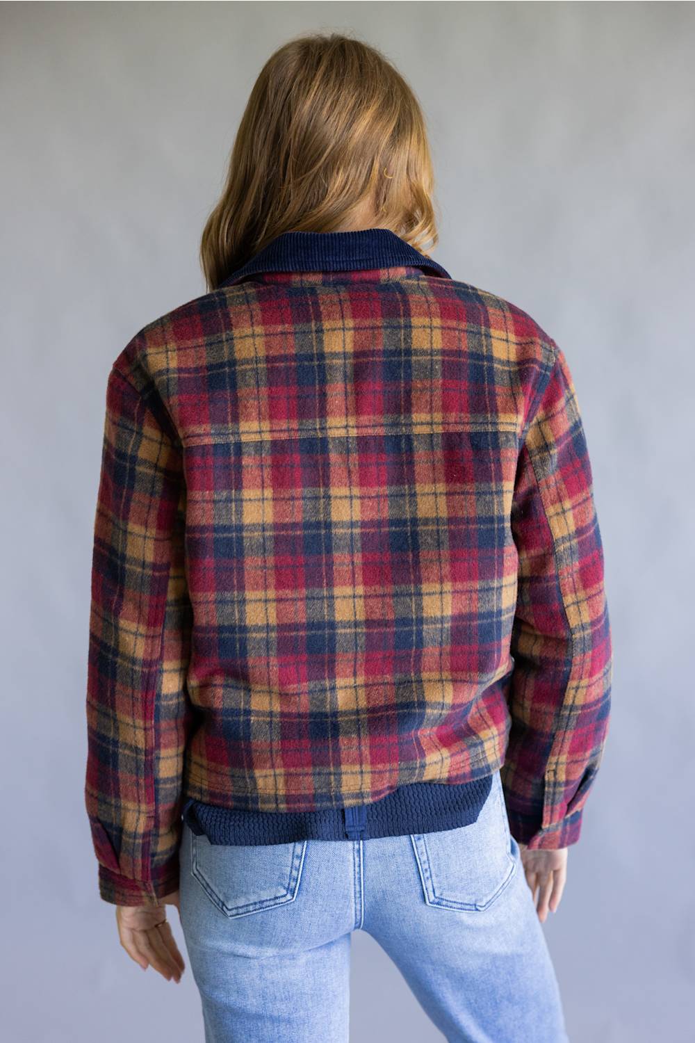 A woman viewed from behind wearing a red, navy, and yellow plaid flannel jacket with a navy ribbed collar, cuffs, and hem, paired with light blue jeans against a neutral gray background.