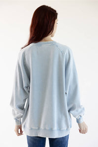A woman viewed from behind wearing a light blue washed crewneck sweatshirt and blue jeans against a plain white background.