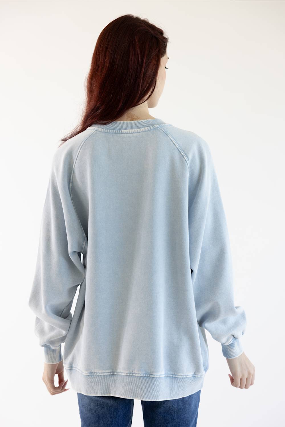 A woman viewed from behind wearing a light blue washed crewneck sweatshirt and blue jeans against a plain white background.