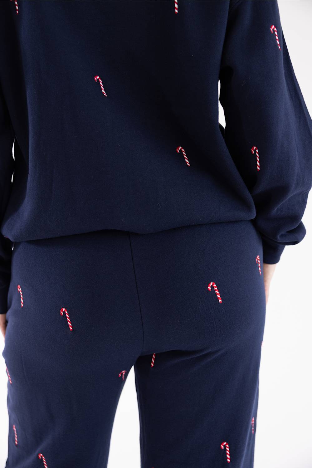 Close-up back view of a woman wearing a navy blue fleece crewneck sweatshirt and matching straight-leg sweatpants, both featuring an all-over embroidered red and white candy cane pattern.