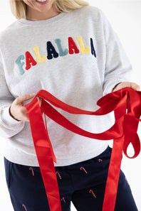 Studio portrait of a blonde woman wearing an 1897 Active Falalala Patch Embroidered Fleece Sweatshirt for Women in Ice Grey with colorful chenille lettering and dark blue holiday-patterned lounge pants.