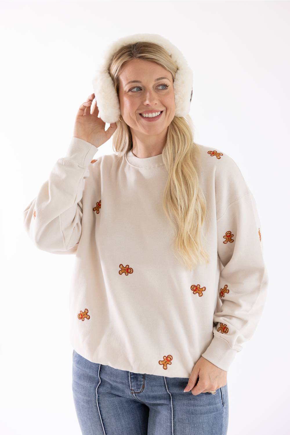 A smiling blond woman wearing fluffy white earmuffs and a beige fleece crewneck sweatshirt with an all-over embroidered gingerbread man pattern.