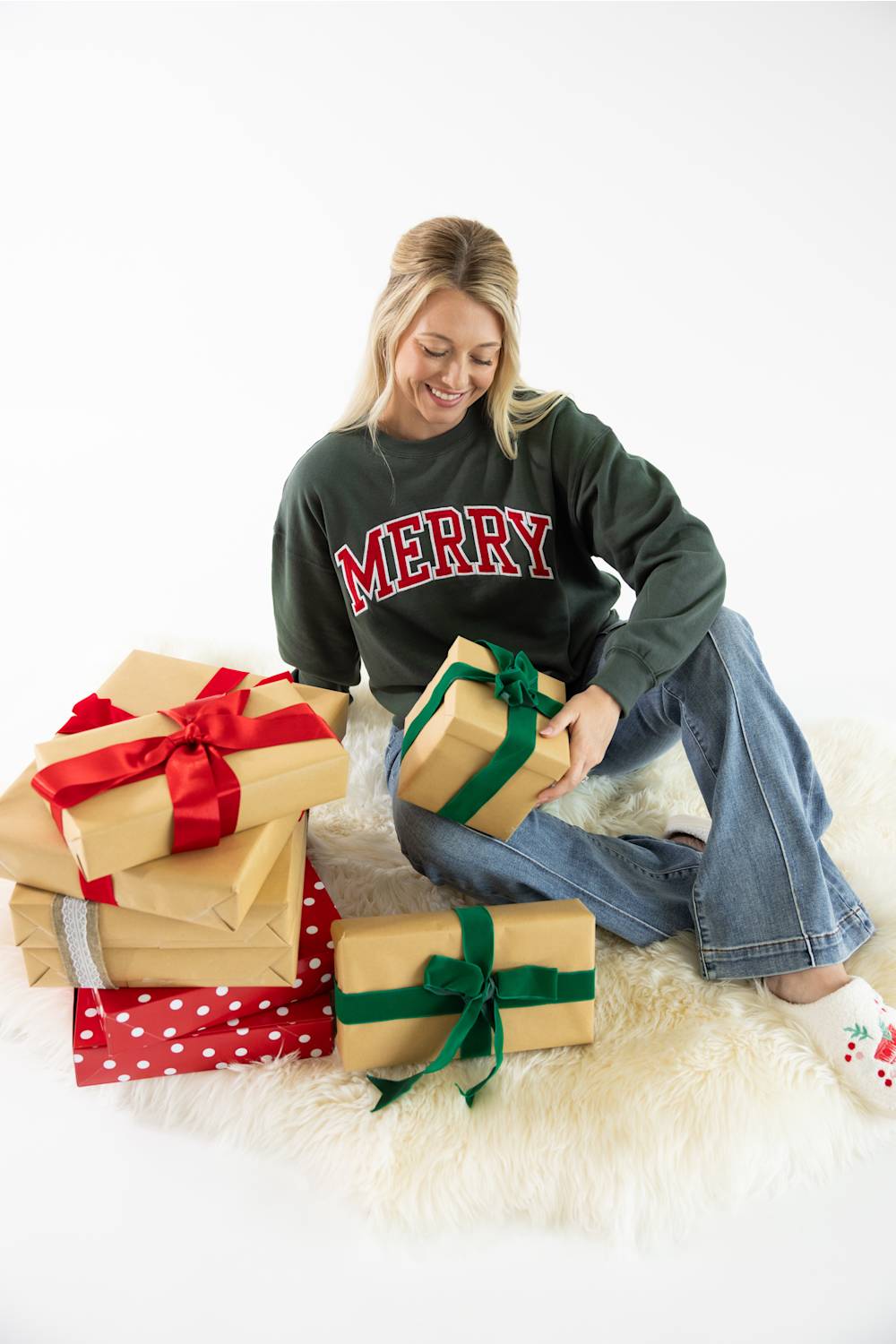 A smiling blond woman sitting on a white faux fur rug next to wrapped presents and a Christmas tree, wearing a dark green crewneck fleece sweatshirt featuring red and white block lettering that reads "Merry" across the chest, light wash jeans, and holiday-themed slippers.