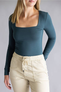 A woman wearing a fitted long-sleeve top with a square neckline in a muted teal-blue color, paired with light beige drawstring pants, against a plain light gray studio background.