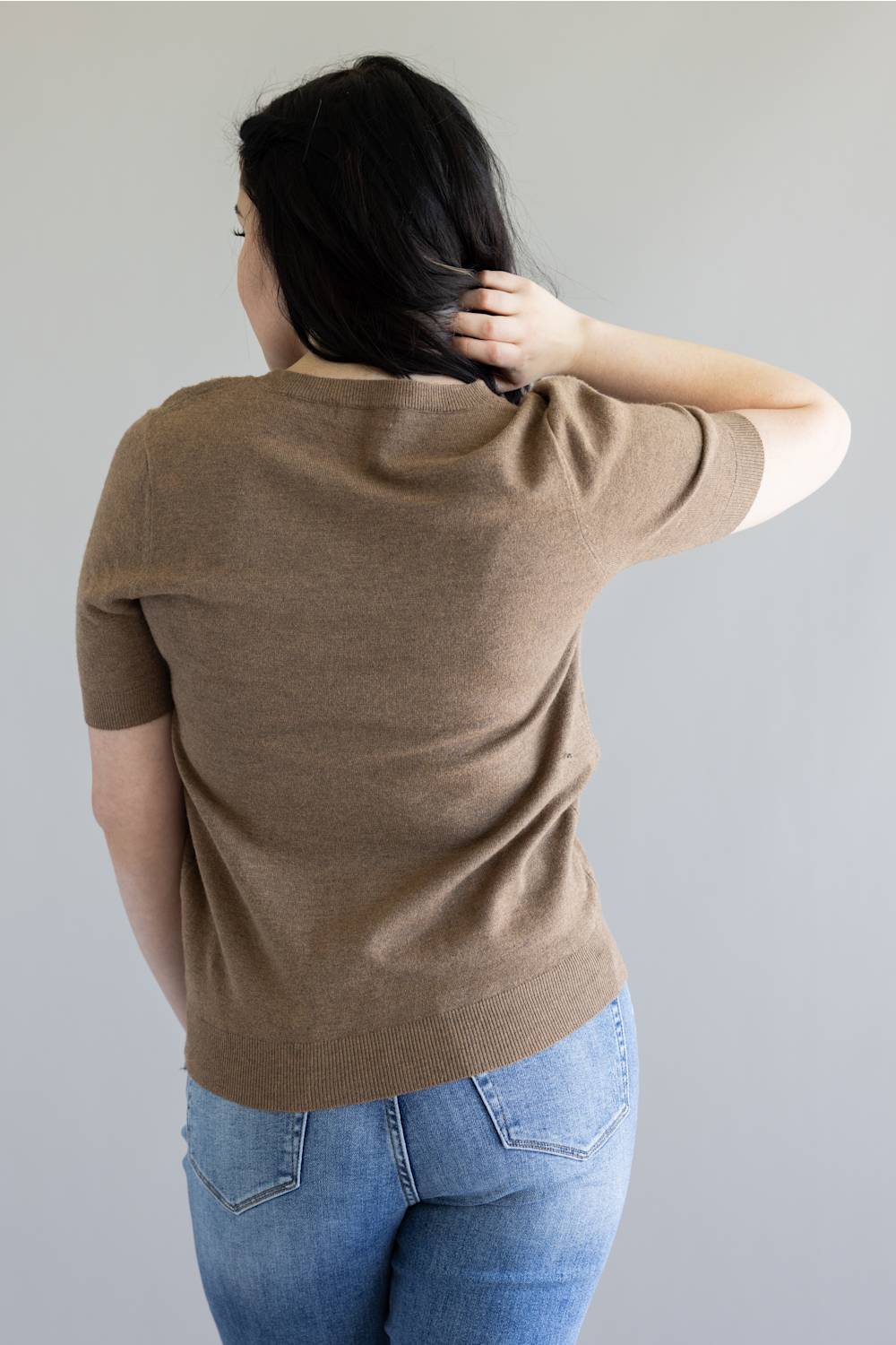 A woman shown from behind wearing a short-sleeve knit top in a warm taupe-brown color, paired with blue jeans, against a plain light gray background.