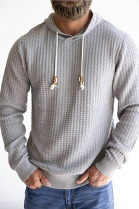 A man wearing a light gray waffle-knit hoodie with white drawstrings and blue jeans, shown from the neck down against a plain white background.