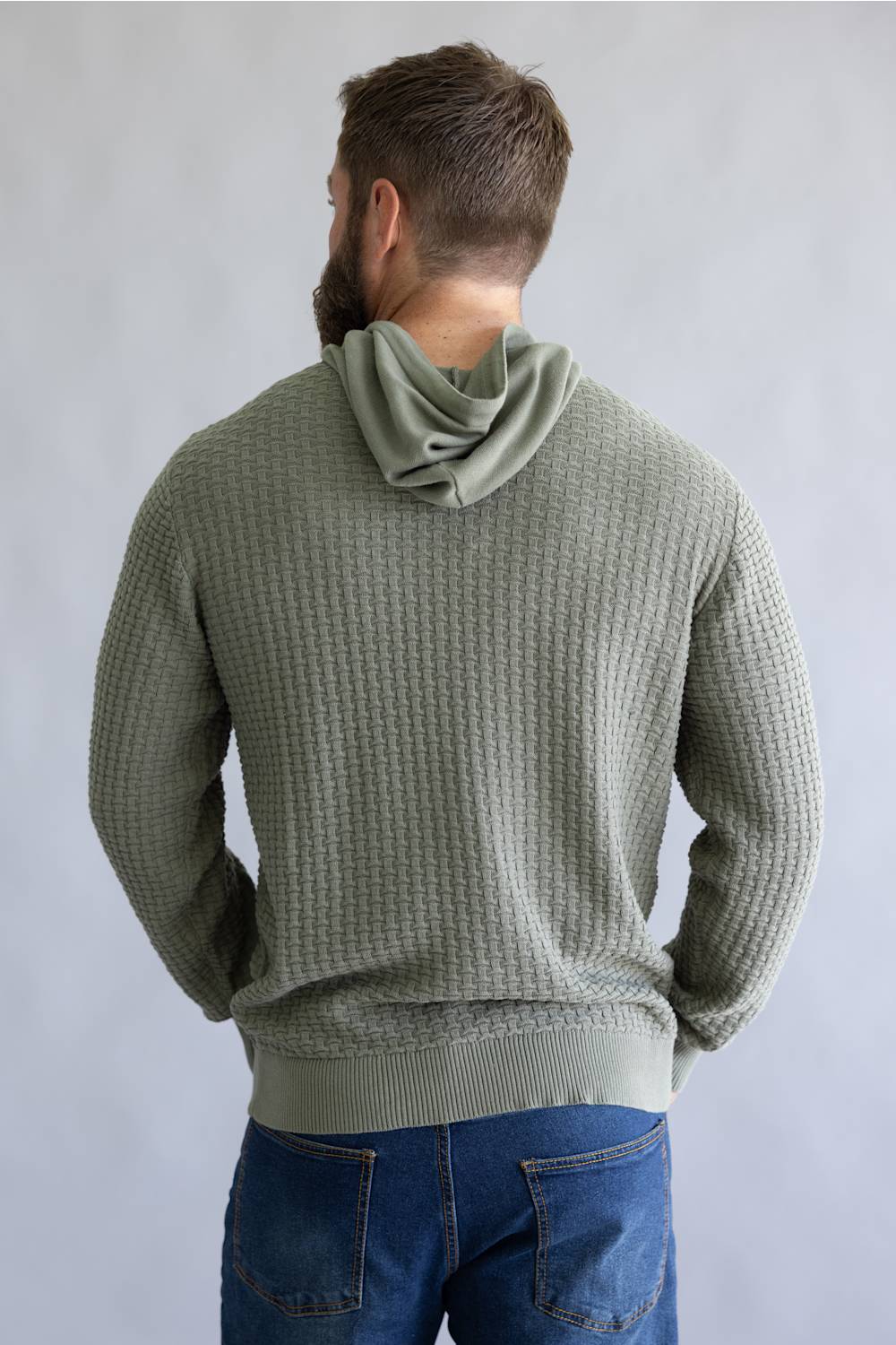 A man viewed from behind wearing a sage green waffle-knit pullover hoodie with a cowl neck, paired with blue jeans, against a neutral gray background.