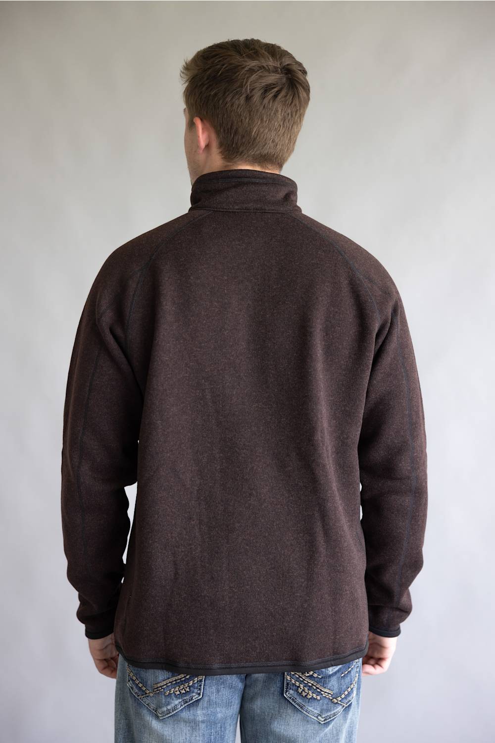 A man viewed from behind wearing a dark brown long-sleeve fleece pullover and blue jeans, standing against a plain light gray studio background.