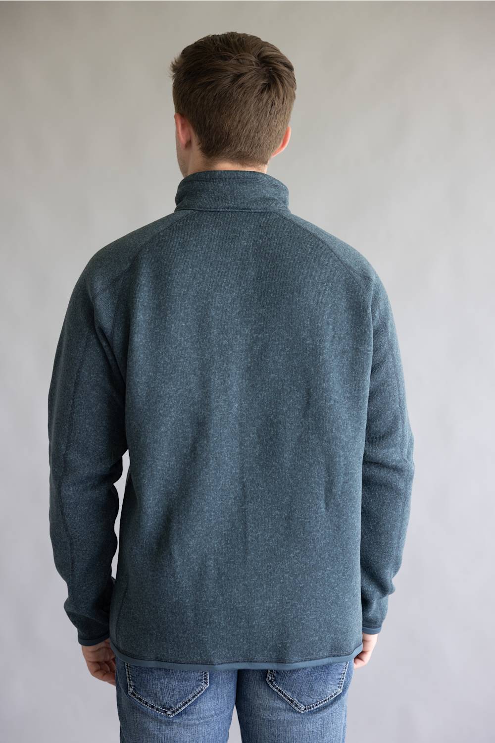 A man viewed from behind wearing a dark slate blue heathered quarter-zip fleece pullover and blue jeans, standing against a neutral light gray studio background.