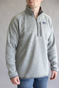 A man wearing a heather gray quarter-zip fleece pullover with a zippered chest pocket and a small Patagonia logo patch on the left chest, paired with blue jeans against a neutral gray background.