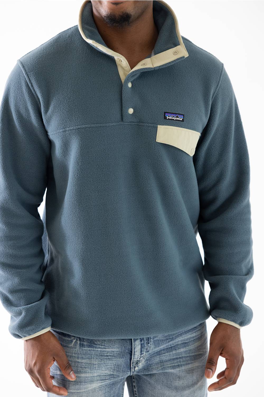 A man wearing a Patagonia fleece pullover in a muted slate blue with beige contrast trim and a chest pocket, paired with blue jeans against a white background.