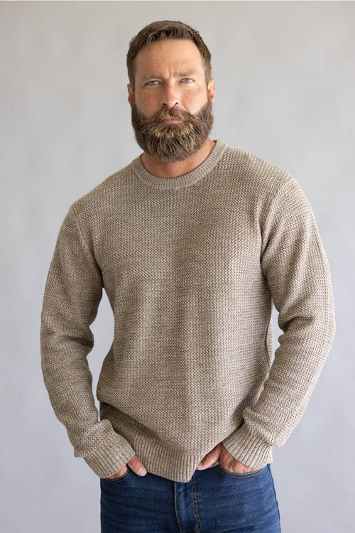 A man with a full beard wearing a textured, ribbed knit crewneck sweater in a warm taupe-beige color, paired with blue jeans, standing against a neutral gray studio background.