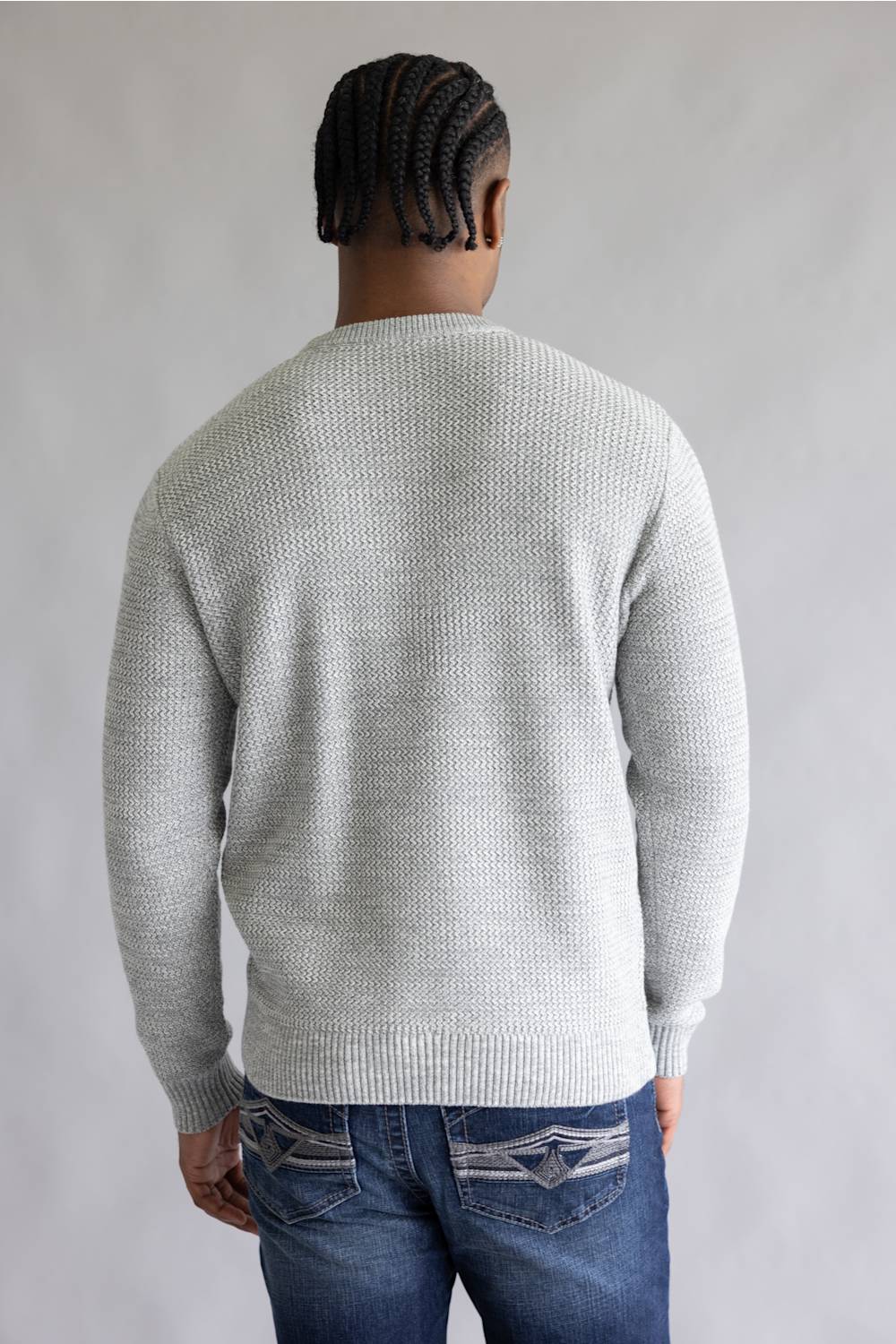 A man viewed from behind wearing a light gray textured knit sweater and blue jeans, standing against a plain light gray background.
