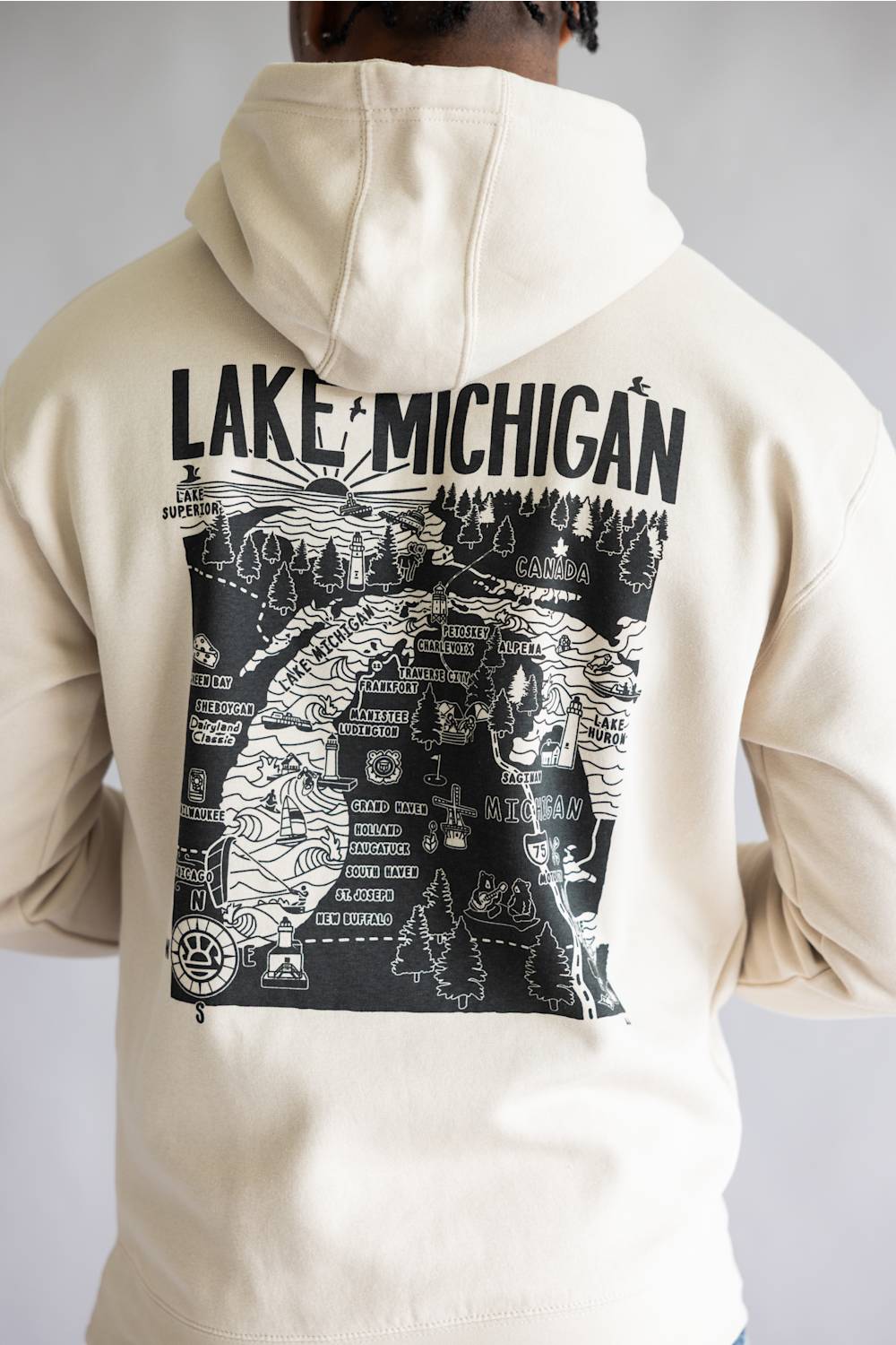 A person shown from the back wearing a cream-colored hoodie with a large black graphic print on the back featuring a map illustration and the text 'LAKE MICHIGAN'.