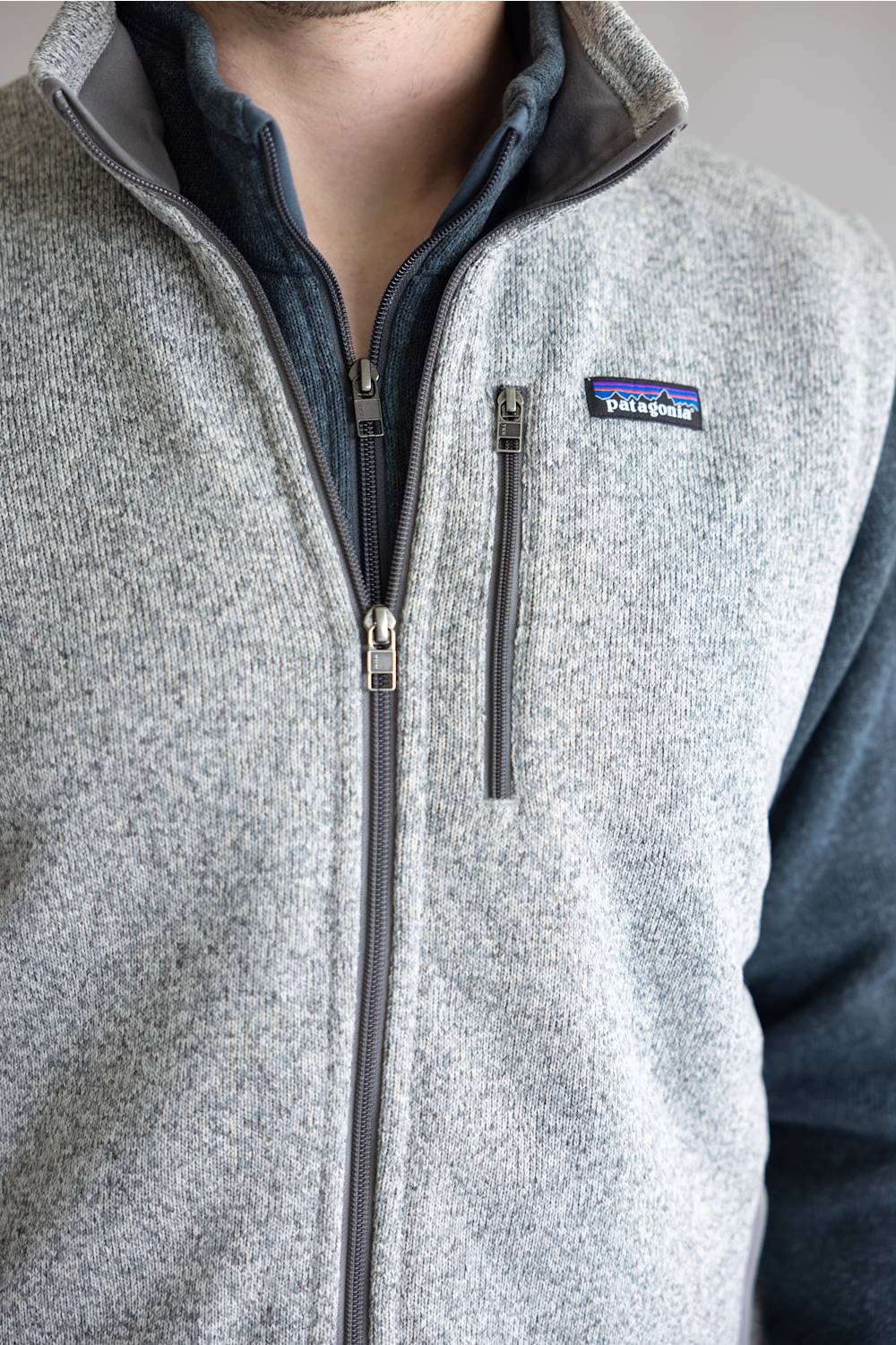 Close-up of a person wearing a heather gray zip-front fleece vest with a dark gray inner collar and a visible Patagonia logo patch on the chest.