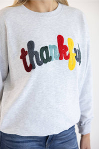 A person wearing a light heather gray crewneck sweatshirt with the word 'thankful' spelled out in colorful raised letters across the chest, paired with blue jeans.