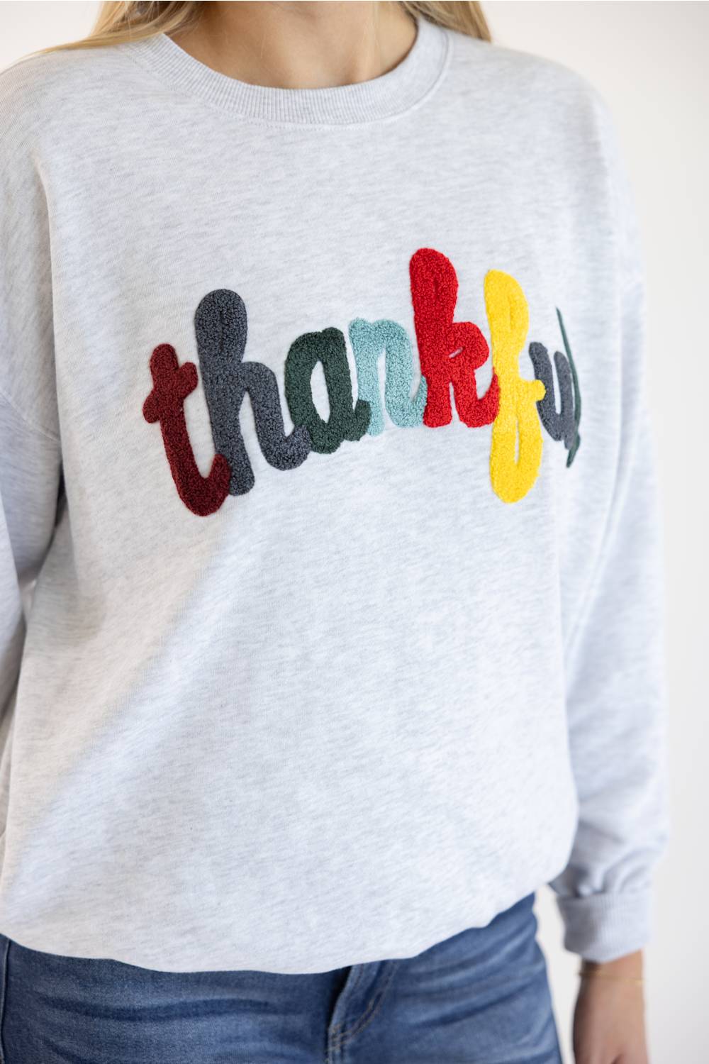 A person wearing a light heather gray crewneck sweatshirt with the word 'thankful' spelled out in colorful raised letters across the chest, paired with blue jeans.
