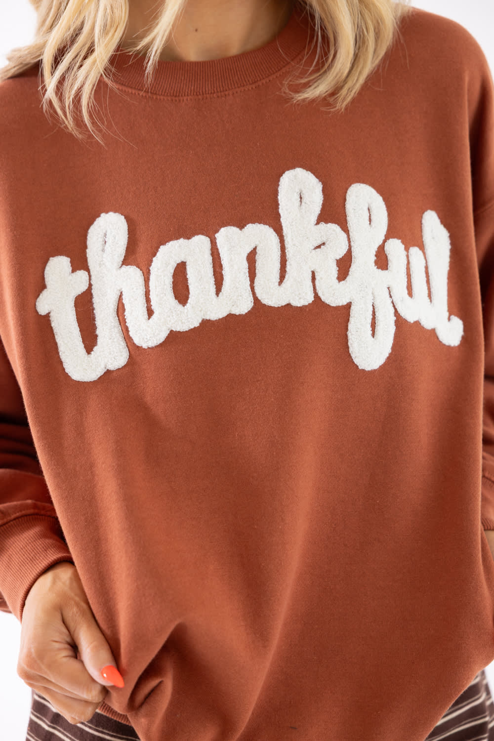 A close-up of a person wearing a rust-brown crewneck sweatshirt with the word 'thankful' in large white fuzzy cursive letters across the chest.