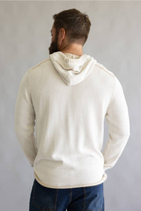 A man viewed from behind wearing a cream-colored knit hoodie with decorative stitching along the shoulders, paired with dark blue jeans, against a neutral gray background.