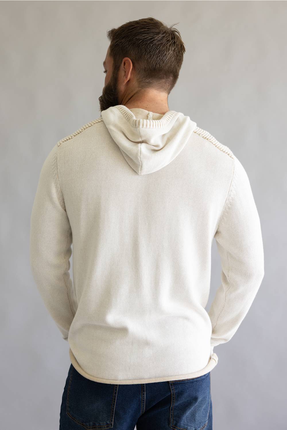A man viewed from behind wearing a cream-colored knit hoodie with decorative stitching along the shoulders, paired with dark blue jeans, against a neutral gray background.