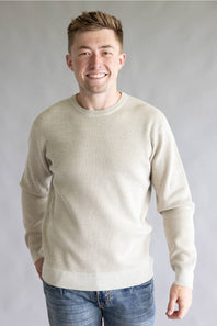 A smiling man wearing a light beige crewneck knit sweater and blue jeans, standing against a neutral gray studio background.