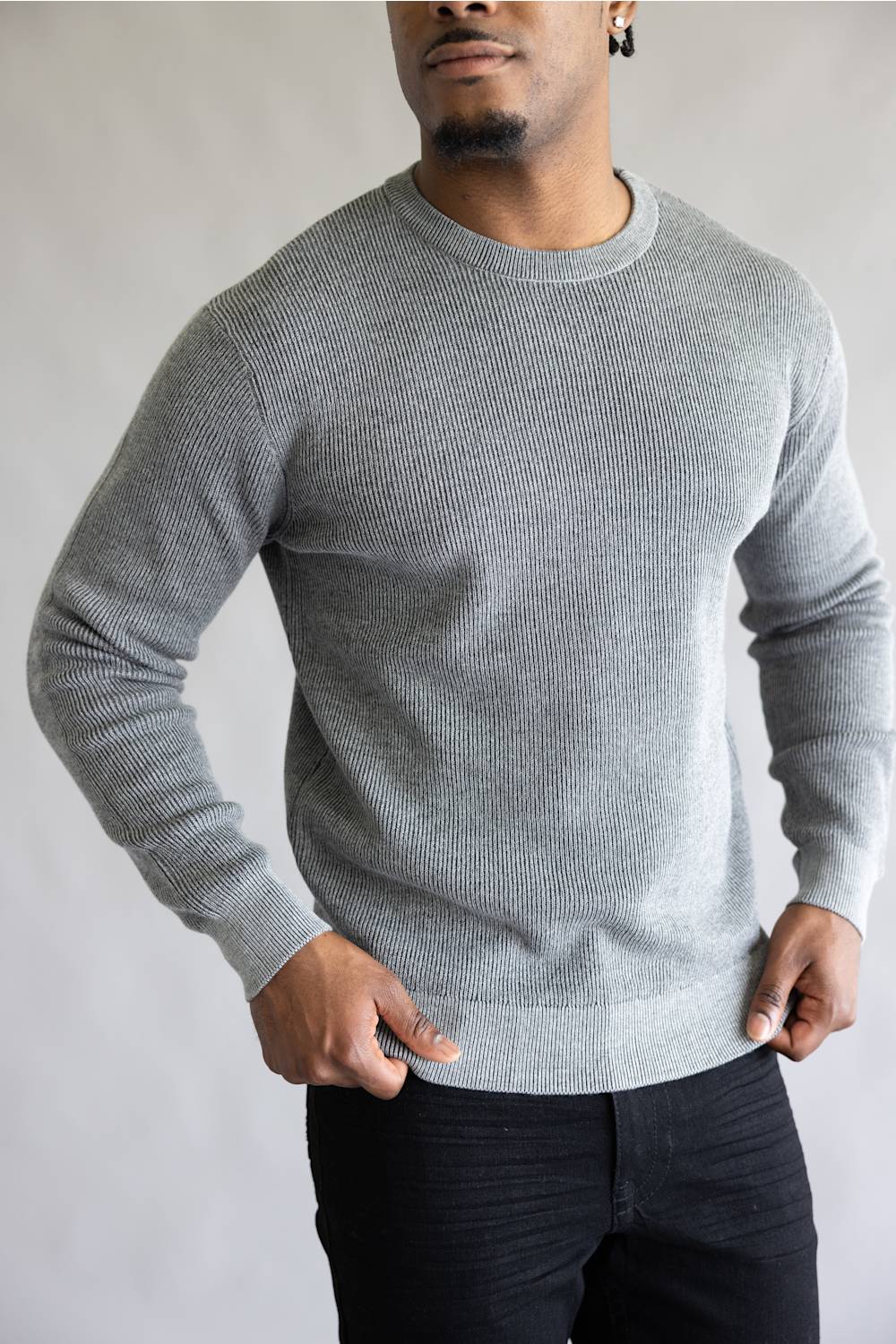 A man wearing a light gray ribbed knit crewneck sweater and black pants, standing against a neutral gray background.
